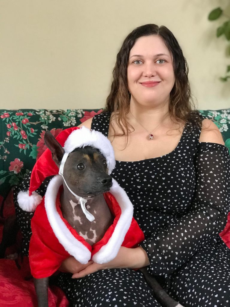Photo of a pretty young woman with light brown wavy hair, wearing a black dress with tiny white hearts and a red "Russian Faberge egg" necklace. She is holding a dark brown hairless dog with dark eyes, pointy ears, and a white speckled chest. The dog is wearing a Santa suit and hat.