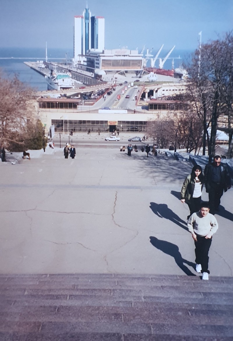 The background is a photo of the Port of Odessa — a massive structure with numerous buildings, including the multistory Hotel Odessa, and dock areas. One large ship is visible at the dock. There are cars, trees, and pedestrians in view. The foreground shows a male and female couple and a teenage male approaching a huge stone staircase.