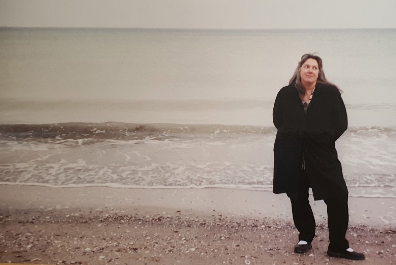 A middle age woman with long, windblown salt and pepper hair wearing a heavy black coat and black pants is standing awkwardly on the shore of a gloomy beach with an overcast sky.