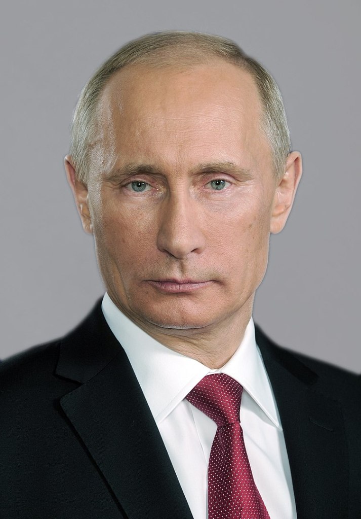 Photograph of Vladimir Putin wearing a black suit jacket, a white shirt, and a red tie with tiny white dots.
