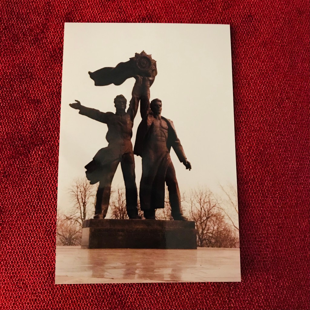 The photo is of two men standing together and holding up a giant medallion with a large ribbon waving toward the left. There are bare trees in the bottom of the background.