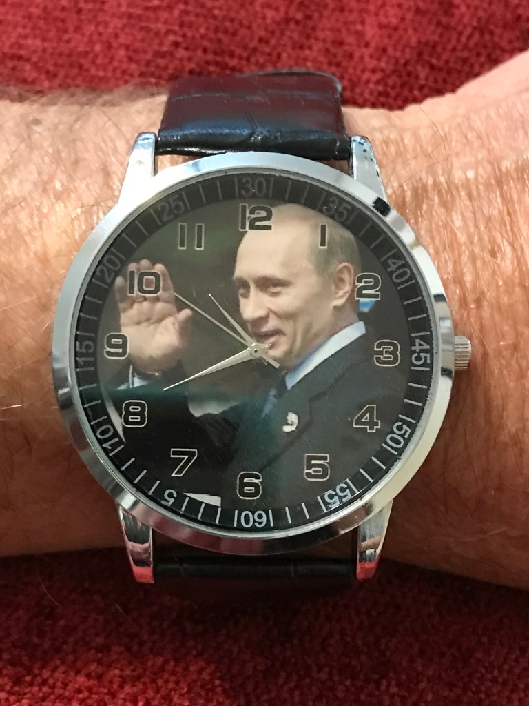Photo is of a watch with Vladimir Putin waving on the face. the watch is on a hairy arm.