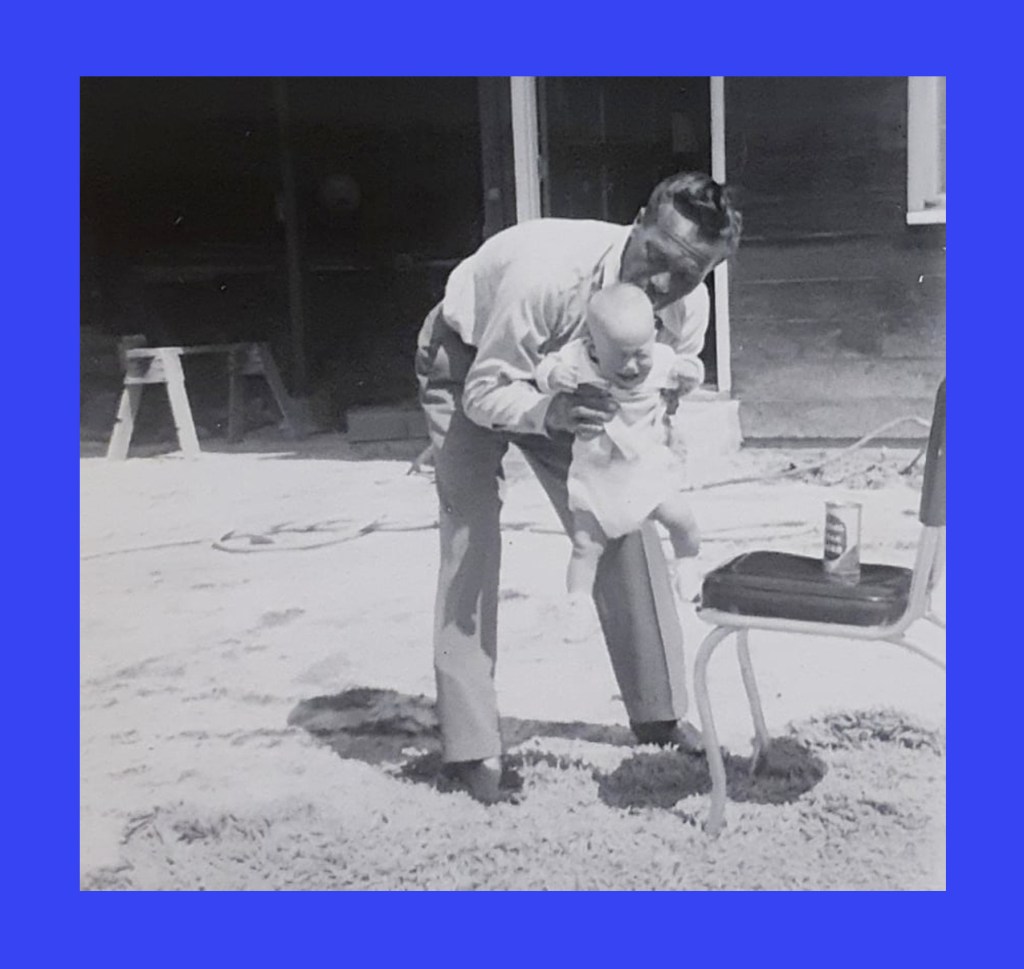Black and white photo of a middle-age man with a dark complexion and dark hair bent over and holding an infant with a fair complexion. The man is wearing tan or light brown slacks and a long-sleeve white dress-shirt. The baby is wearing a white shirt, a diaper, and white shoes. The baby is crying. Nearby is a chair with a beer can on the seat. A sawhorse is visible in the background.
