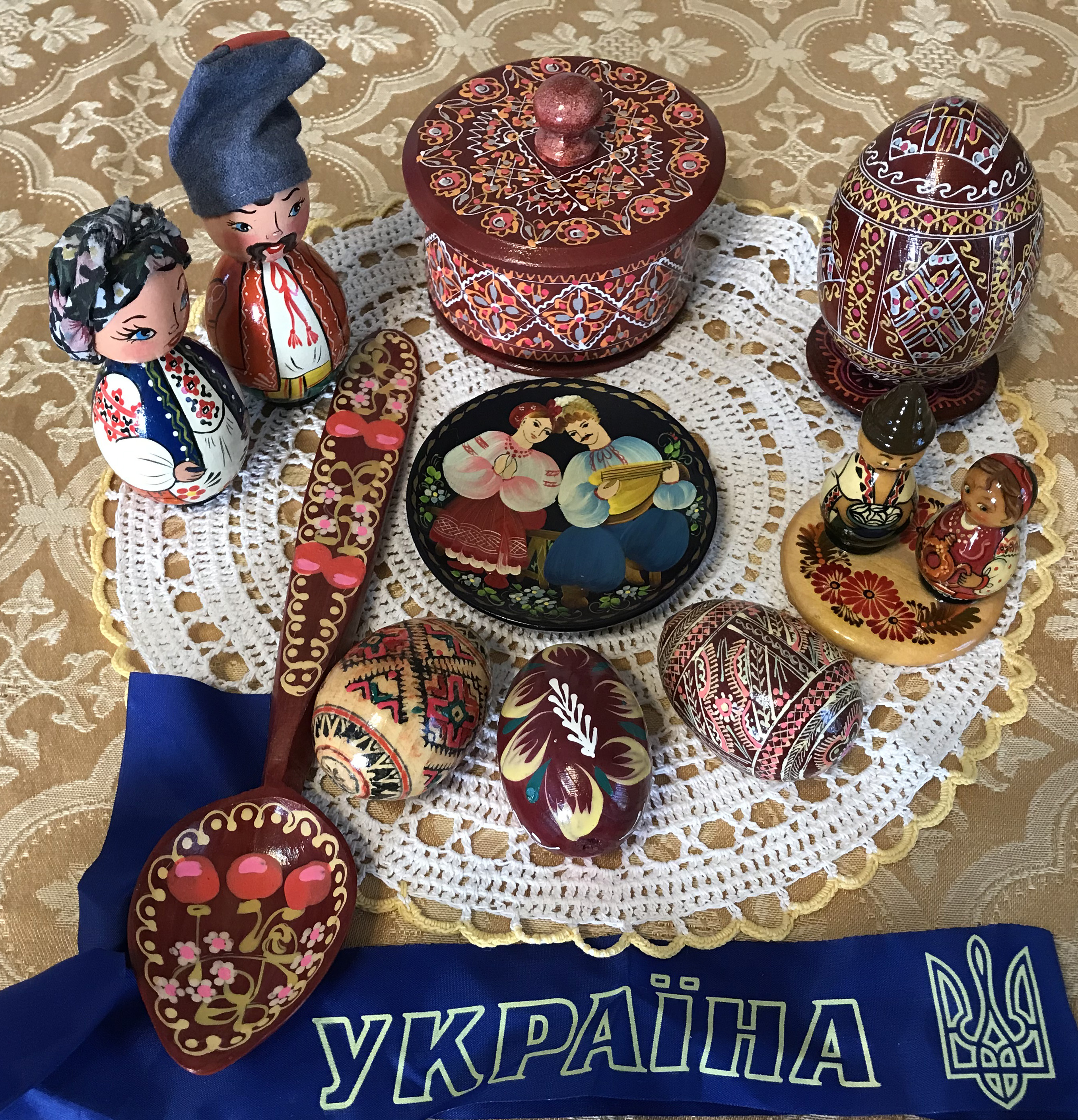 Photo is of an assortment of souvenirs from Ukraine on a white doily on a gold tablecloth. There is a blue ribbon with YKPAIHA (Ukraine) and the Ukrainian lesser coat of arms written on it in yellow. All of the items are painted wooden objects: a female and a male figurine, a round container with a lid, a large egg, a smaller male and female figurine, three eggs, a spoon, and a small dish with a female and male peasant. The eggs, container, and spoon are red with detailed patterns, and the peasants are multicolored.  