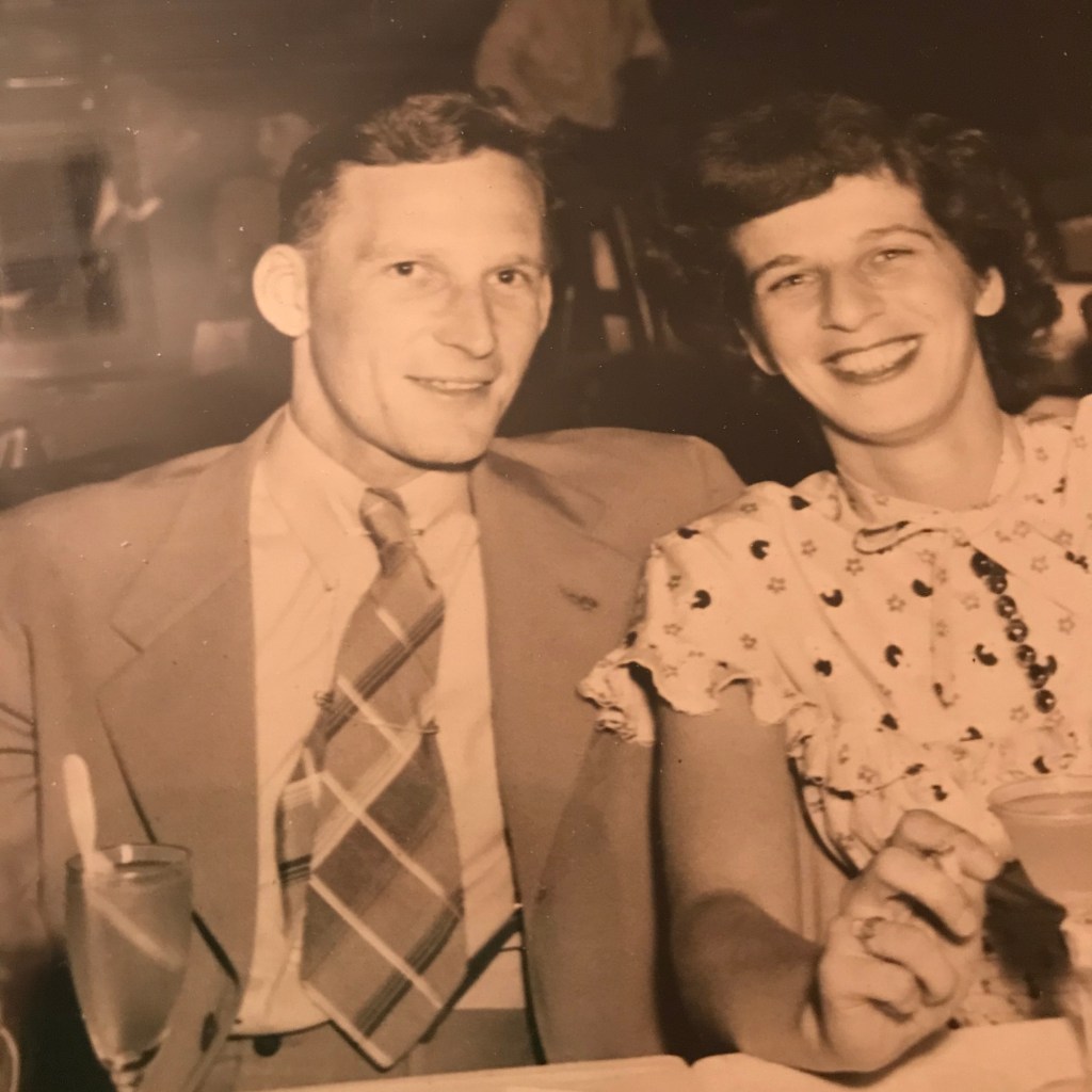 Sepia-toned photo of a man in a suit and a woman with short, dark, curly hair hair. It is the 1950s, and they are seated in a nightclub with cocktails. They are smiling.