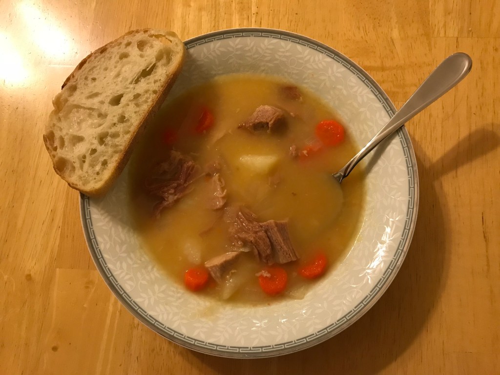 Photo of a bowl of soup with carrots, potatoes, and ham chunks in a yellow broth. The white and silver bowl is on a wooden table. There is a spoon in the soup and a piece of bread resting on the rim of the bowl.