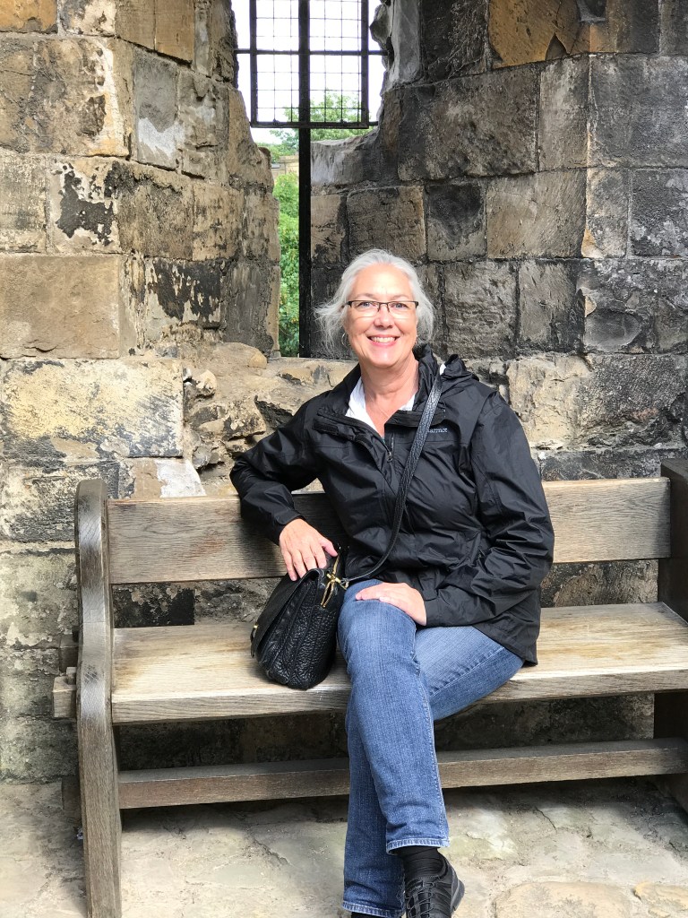 Photo of a gray-haired woman sitting on a bench in front of an old stone building. She is wearing glasses and smiling. She has on blue jeans, a black jacket, and black shoes. Her black purse is next to her. 